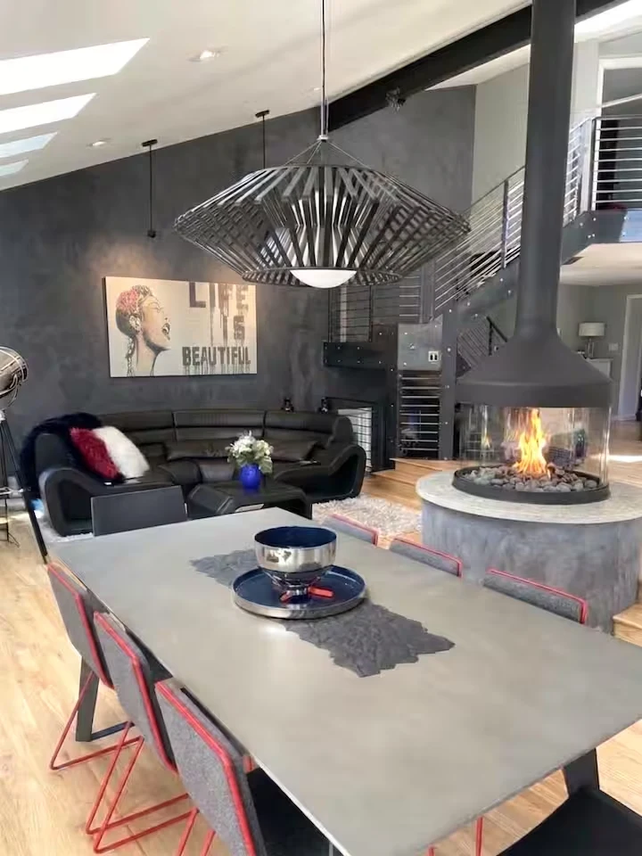 Dining table with red-framed chairs beside the lit circular fireplace, curved sofa and steel staircase