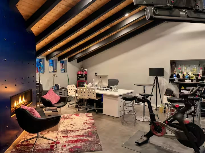 Office with vaulted timber and steel beams, desk, fireplace sitting area, bar and Peloton bike